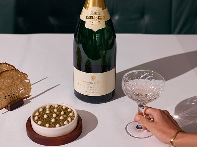 A bottle of Justerini & Brooks Champagne on a dinner table and a person holding a glass with food beside it, including Caviar