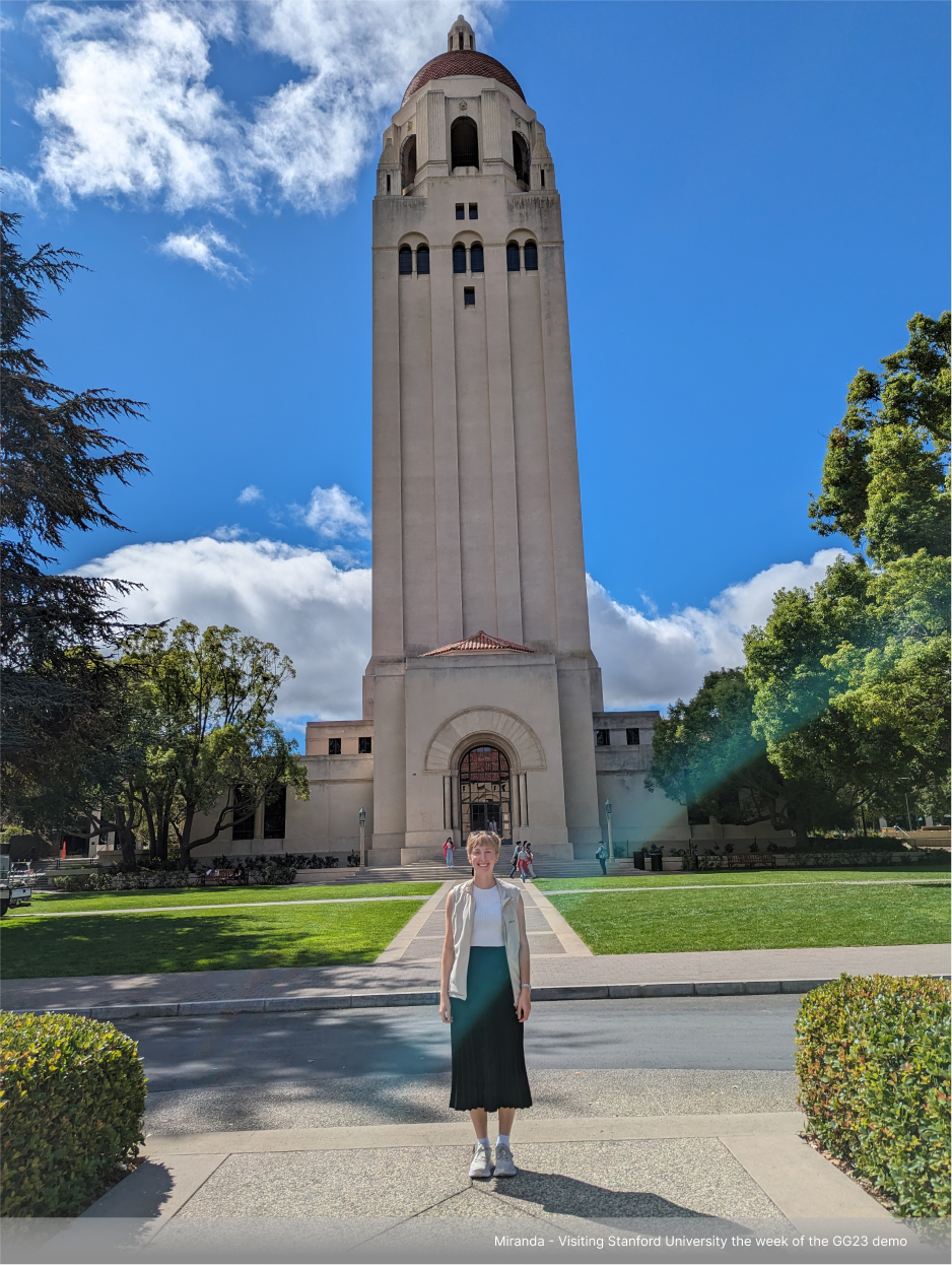 Miranda is standing in front of the iconic Hoover Tower at Stanford University. She is dressed in a white shirt and black skirt, and the sky is blue with scattered clouds. Trees and greenery surround the area, creating a vibrant scene.