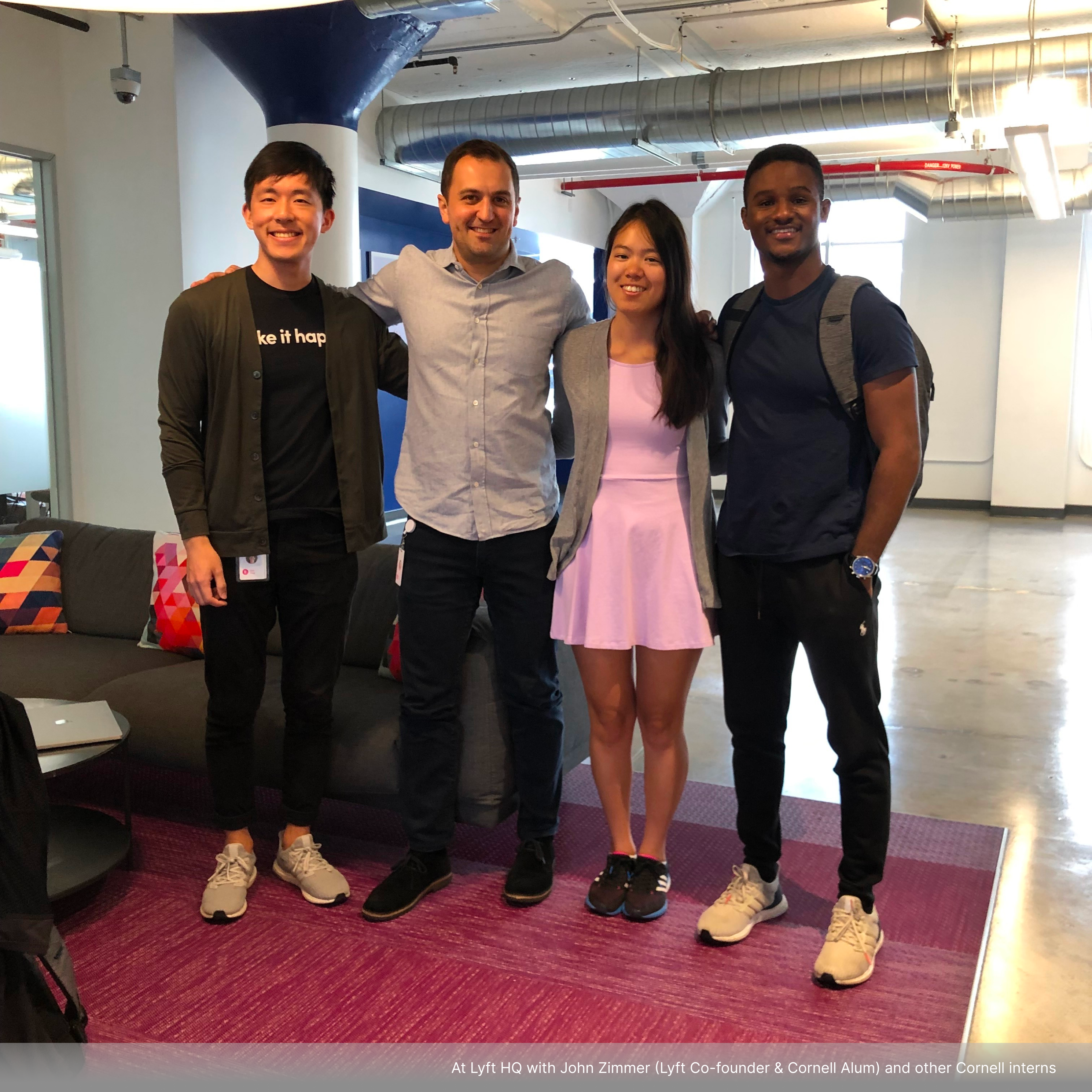 Jasmine standing close together with three others and smiling in a modern office space. Three of them are wearing casual attire while the fourth, in the middle, is dressed in business casual. The group stands in front of a couch with colorful cushions and a pink rug beneath them.