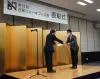 A man in a dark suit presents an award certificate to another man who is COO Hiroshi Mushigami in a grey jacket and red ribbon on stage. A banner overhead reads "The 33rd Nikkei New Office Award Ceremony" in Japanese. A microphone stands nearby.