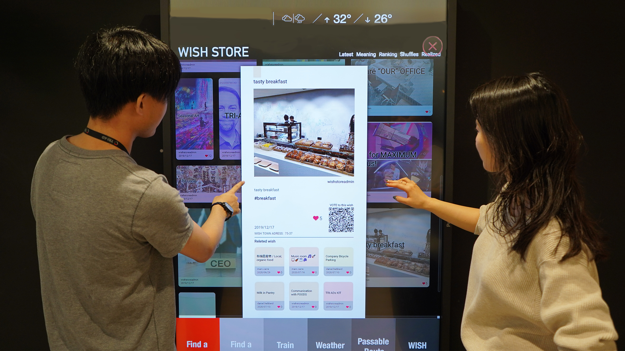 Two TRI-AD employees are interacting with a large touchscreen display labeled "Wish Store" in the office. They are pointing at a digital profile of a bakery with an image of pastries. The screen also shows buttons for "Find a Train," "Weather," and other services.