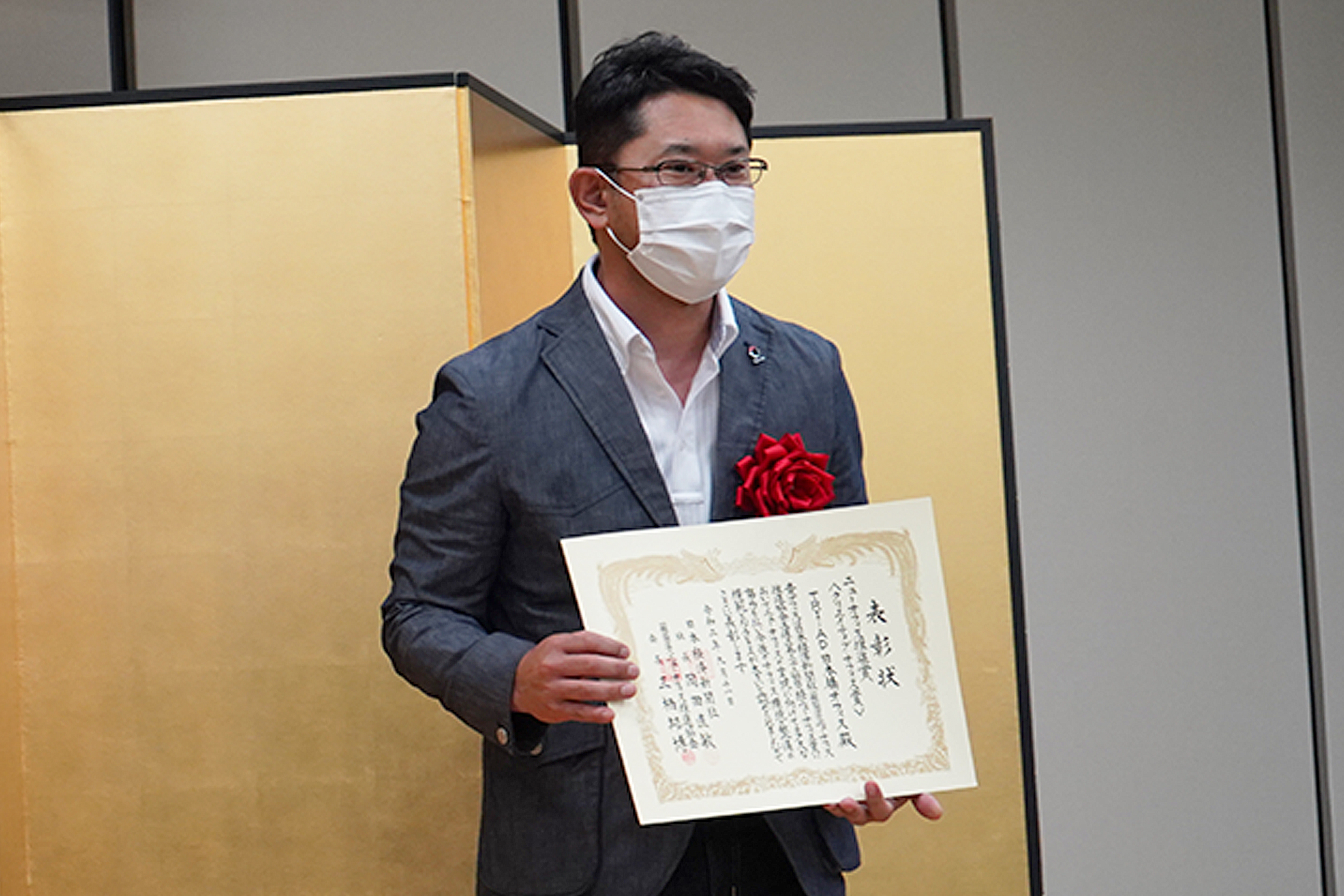 COO Hiroshi Mushigami wearing a mask, glasses, and a gray suit jacket adorned with a red red ribbon on the lapel holds the Creative Office Award. He stands in front of a gold folding screen in a formal setting.　