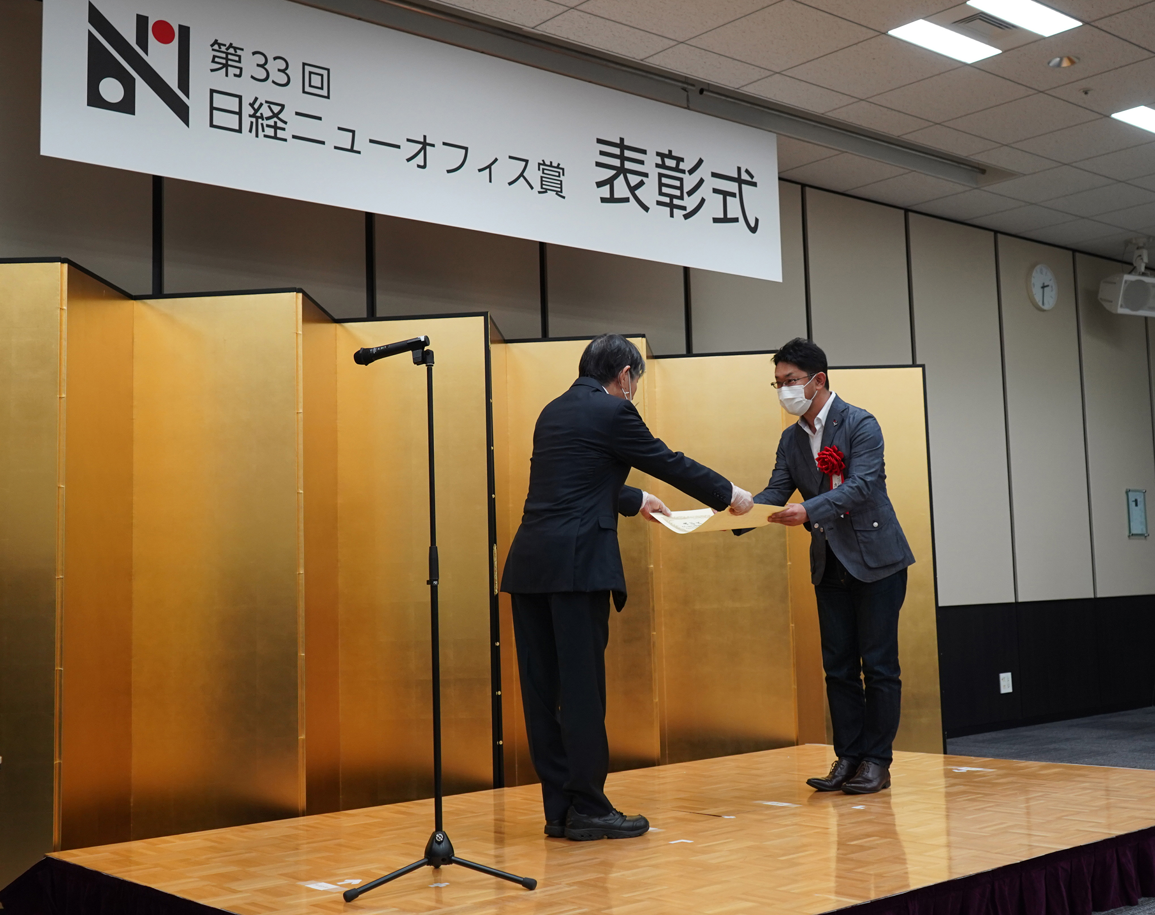 Two men are on a stage during an award ceremony. A man, wearing a suit and mask, is handing a certificate to the other man who is COO Hiroshi Mushigami, also in a suit and mask. A microphone stand is positioned on the stage, and a banner above reads "The 33rd Nikkei New Office Award Ceremony" in Japanese.