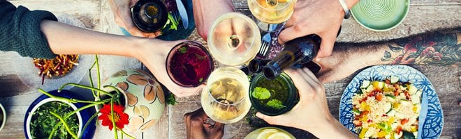 People clinking their drinks glasses over a table laid out with plates and bowls of food.