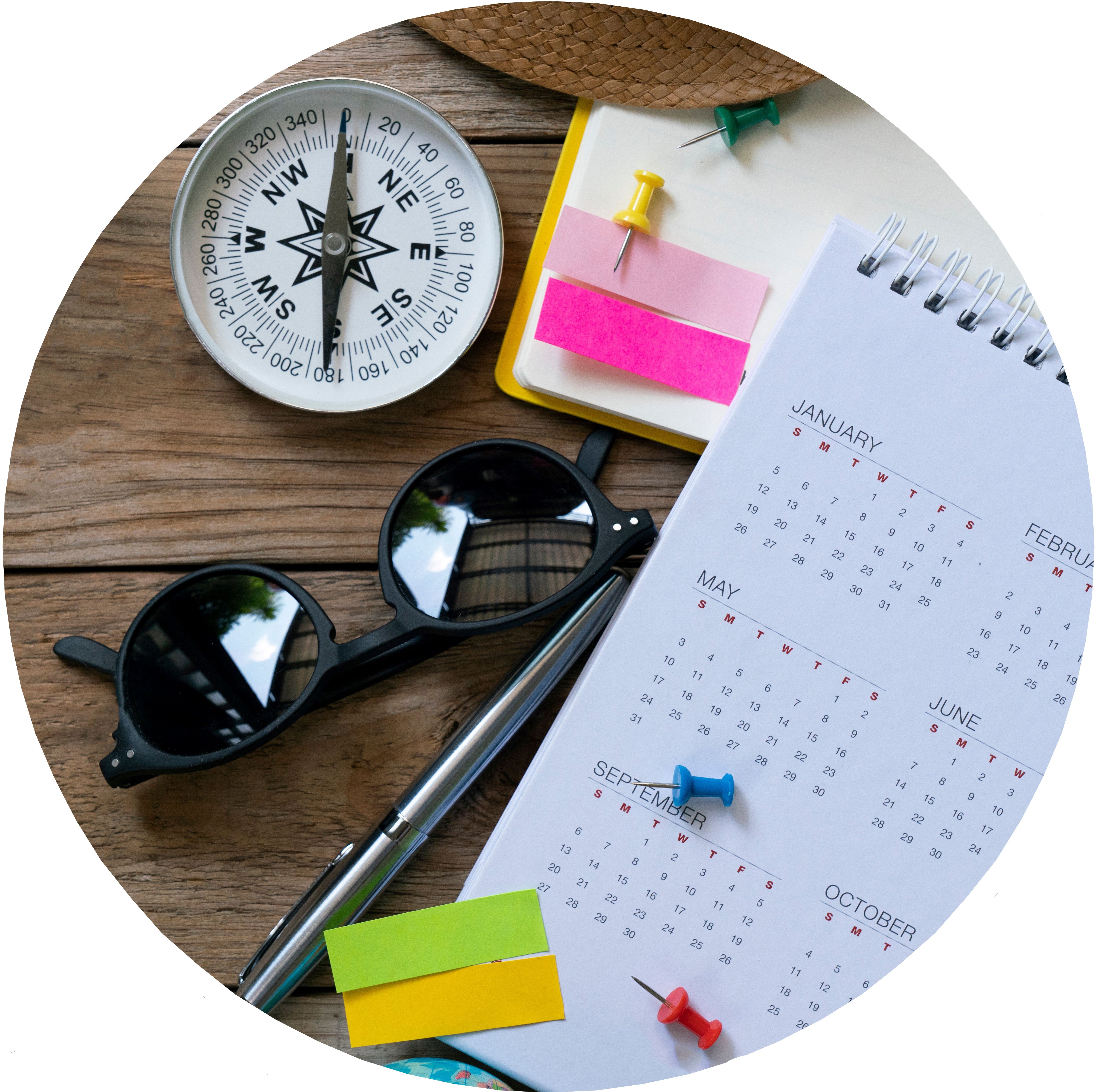 Items bunched on a wooden desk including a compass, sunglasses, a pen, a calendar, pins, sticky notes and a straw hat.