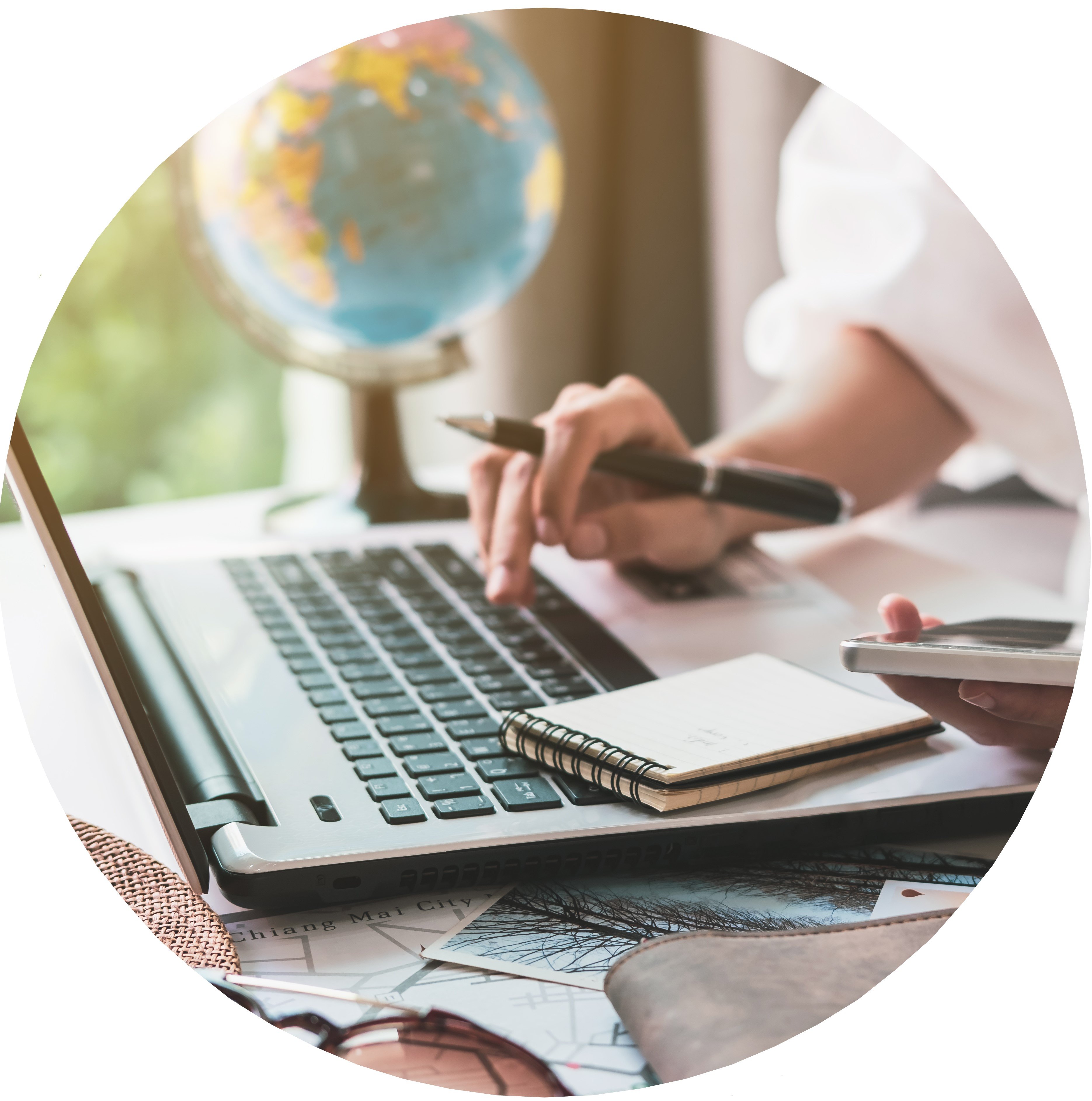 Someone is planning a trip on paper using a laptop at a desk with a globe beside them.