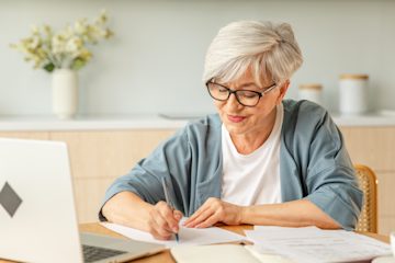 Older woman sitting at a table going through paperwork with her laptop in front of her. She is slightly smiling as she writes.