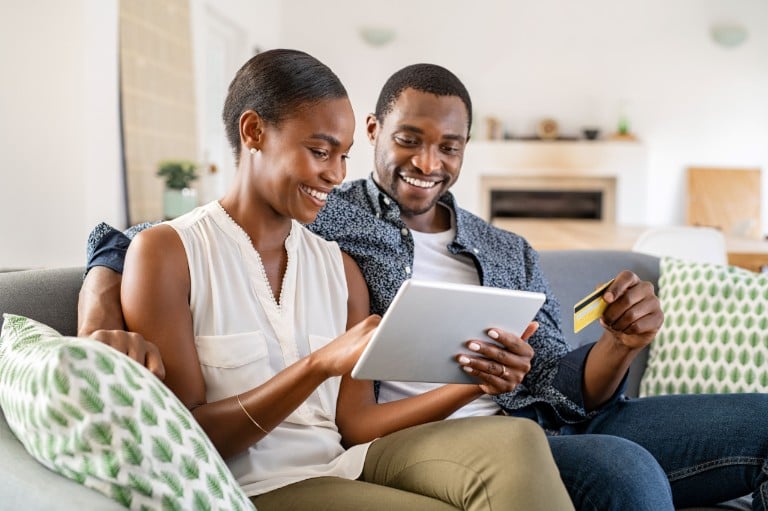 A black man and woman sit on a sofa, smiling at a tablet. The man holds a yellow payment card.