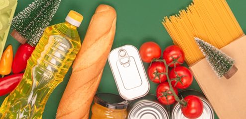 Food laid across a dark green background, including a baguette, a bottle of oil, tomatoes and spaghetti