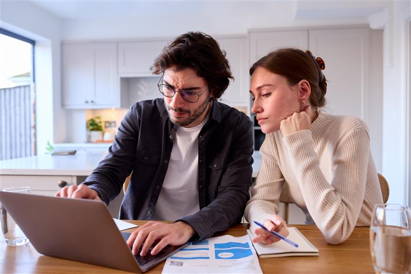 A young couple sitting at a dining table and looking at a laptop, with an energy bill and notebook spread out in front of them