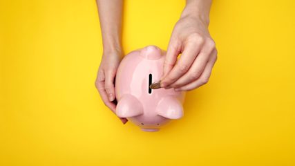 Person putting a coin into a pink piggybank on a yellow background.