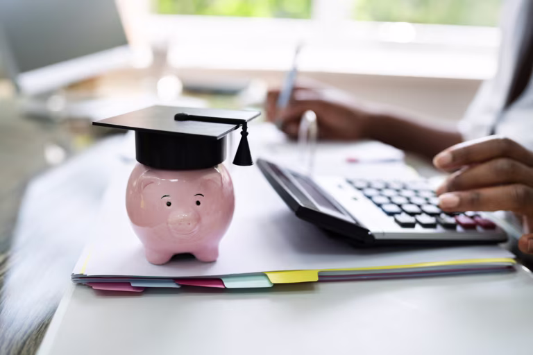 A piggy bank with a graduate cap on a desk next to a calculator