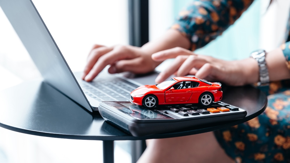 Image of woman typing on laptop with a calculator next to her and a red miniature car toy on top of that. 