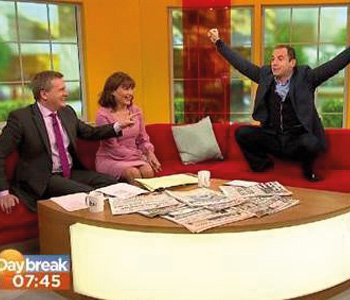 Martin Lewis with his arms in the air kneeling on the Good morning Britain sofa ready to jump off