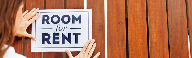 A Caucasian brunette woman puts up a 'ROOM for RENT' sign on a wooden slatted wall.