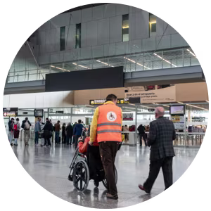 A wheelchair user is being helped through an airport by a member of staff wearing orange hi-vis.