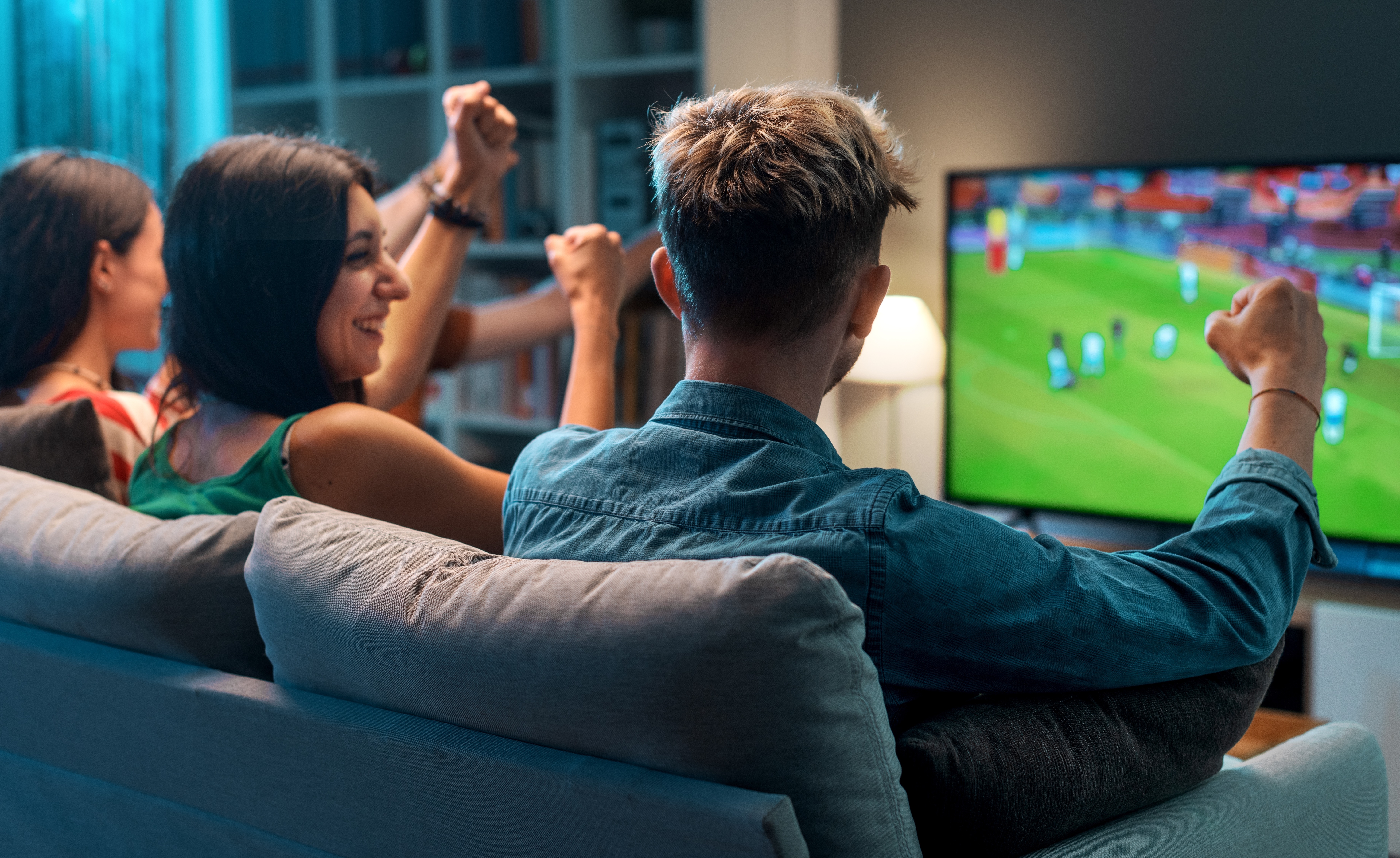 Two women and a man cheering while watching a football match on TV