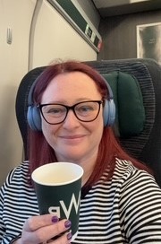 Woman sat on train with a cup of tea wearing headphones. She is in a seat in a first class carriage. She is similing. It's early in the morning so there is no natural light.