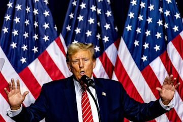 US President Donald Trump giving a speech in front of three American flags.