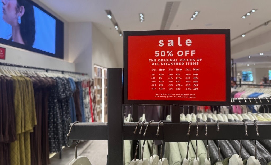 A clothing shop interior with red sign saying "sale 50% OFF", above racks of clothes.