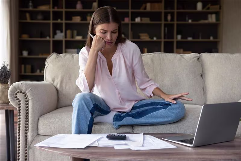 A young woman wearing a pink button-down shirt and jeans sits on a sofa, looking at a laptop and papers on the table in front of her with a frustrated expression on her face