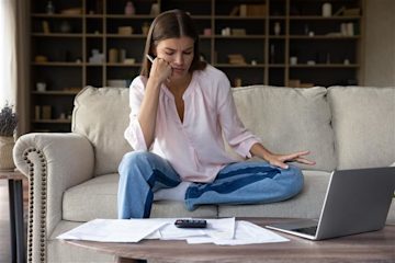 A young woman wearing a pink button-down shirt and jeans sits on a sofa, looking at a laptop and papers on the table in front of her with a frustrated expression on her face