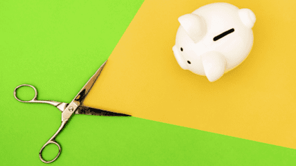 A white piggy bank on a bright green and yellow background, with a pair of scissors coming towards it