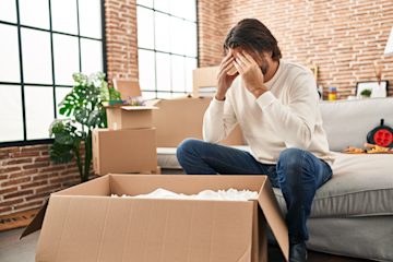 Man in a house sitting on a sofa with his head in his hands. Around him are half-packed cardboard boxes.