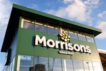 A Morrisons storefront with a large logo and glass windows against a bright blue sky