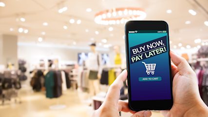 A person in a clothes shop holding a phone with 'buy now, pay later!' written on it.