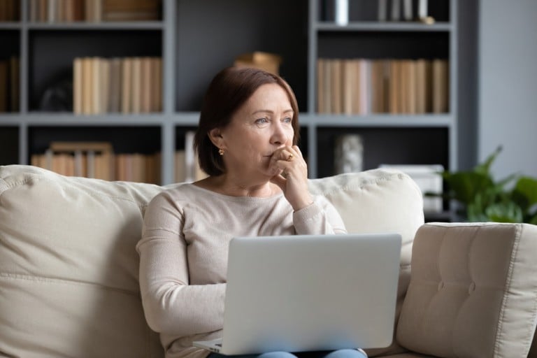 A Caucasian brunette woman sits on a sofa, looking into the distance, with a laptop on her lap.