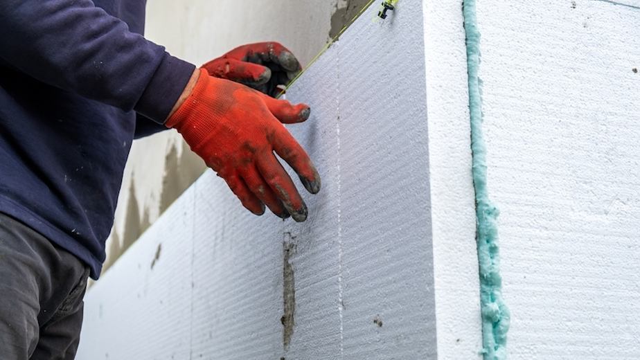 A person wearing orange working gloves installing white insulation on the side of a house