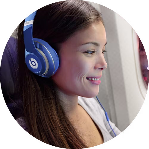 Young woman wearing blue Beats headphones smiling while sitting on an airplane.