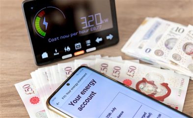 A smart meter, a stack of fifty pound and ten pound notes, and a smartphone with an energy account pulled up spread, across a wooden table.