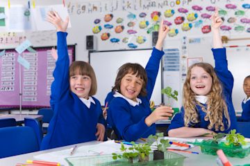 Three primary school children in a classroom with their hands in the air.