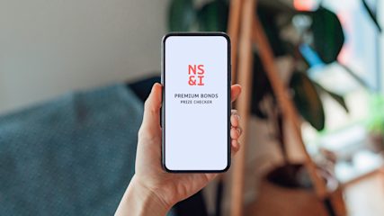 Hand holding a smartphone showing the NS&I Premium Bonds prize checker app