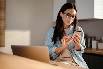 Woman with glasses using smartphone beside open laptop at kitchen table