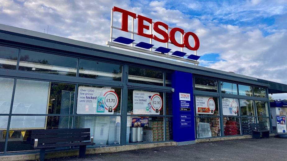 Tesco storefront with a large red-and-blue logo against a bright blue sky with light clouds