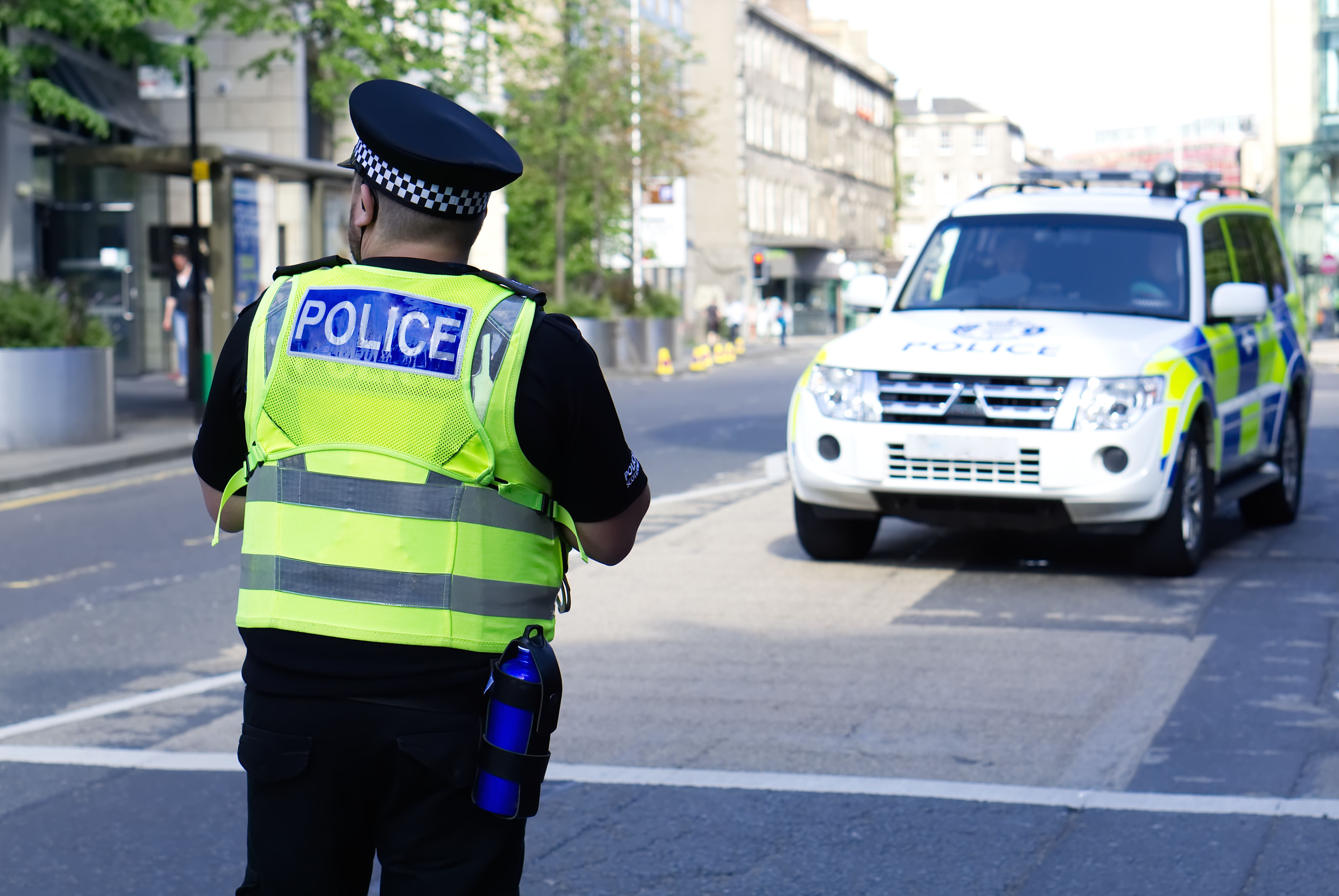 Police officer wearing a high-vis vest with the word "police" on the back, in front of a police car.