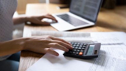 Person at table using a calculator and laptop with paper forms on the table too