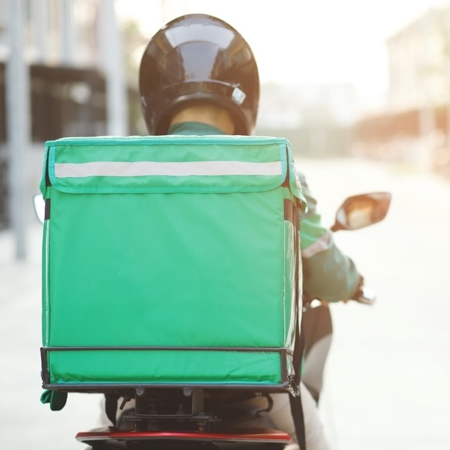 The back of a delivery driver who is riding a motorbike, wearing a black helmet and carrying a green cube backpack.