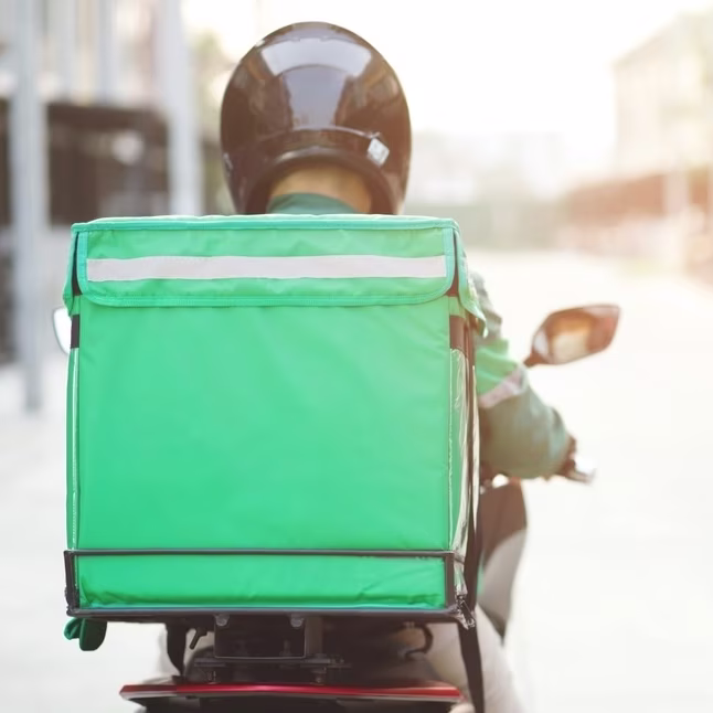 The back of a delivery driver who is riding a motorbike, wearing a black helmet and carrying a green cube backpack.