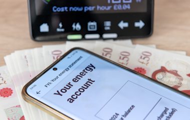 Mobile phone showing 'Your energy account' on the screen on top of a pile of £50 notes, with an in-home device behind.