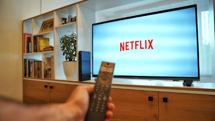 A television set sat on a cabinet in a lounge with the Netflix logo on the screen. A man is holding a TV remote in the foreground.