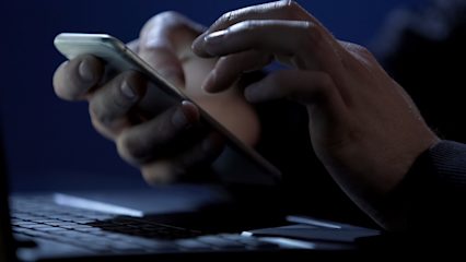 Image shows a person holding a phone in front of a laptop in a dark room