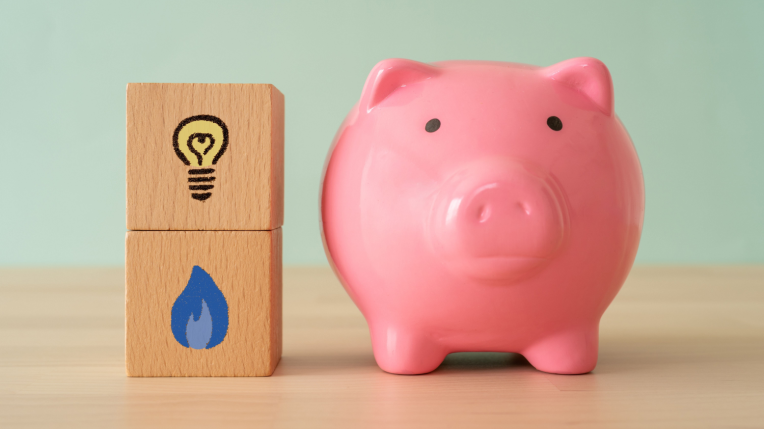 Two wooden blocks are on a table – one with a lightbulb painted on it and one with a gas flame painted on it. Standing next to them is a piggybank.  