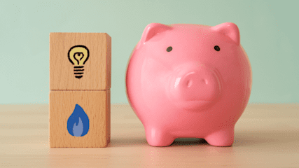 Two wooden blocks are on a table – one with a lightbulb painted on it and one with a gas flame painted on it. Standing next to them is a piggybank.