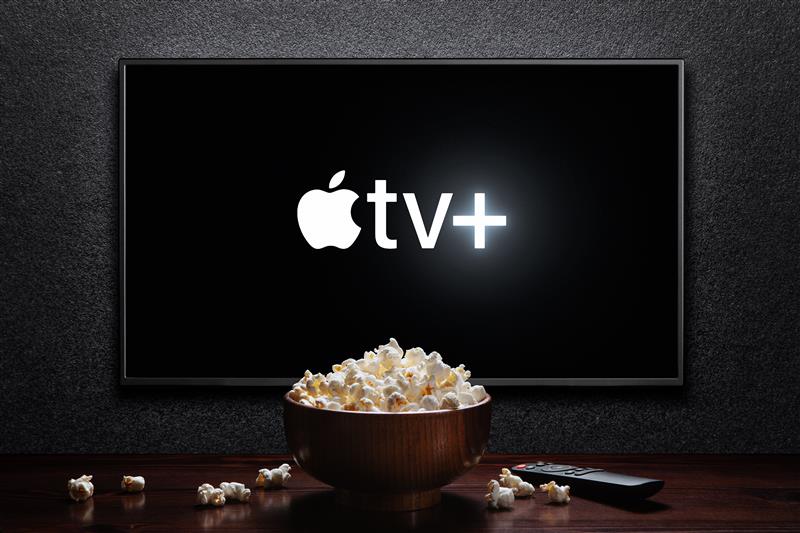 Image shows a TV screen displaying the Apple TV+ logo. In front of the TV screen is a bowl of popcorn on a table.