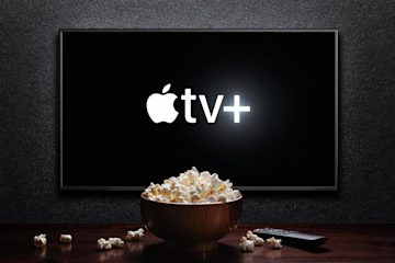 Image shows a TV screen displaying the Apple TV+ logo. In front of the TV screen is a bowl of popcorn on a table.