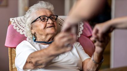 An older woman with white hair and glasses smiles from an armchair while another person reaches toward her, offering assistance.
